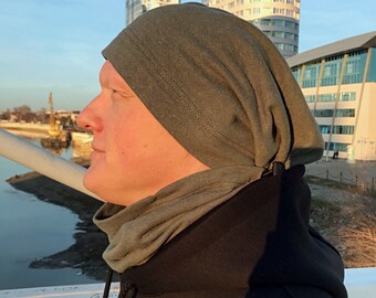 May include: A person wearing a green beanie and a black hooded sweatshirt. The person is standing in front of a building with a glass facade.