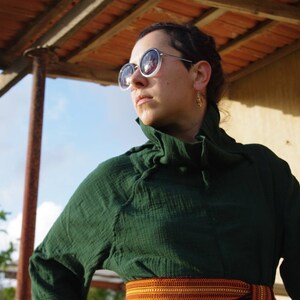 May include: A woman wearing a dark green, collared shirt with a ruffled neckline. She is wearing round, dark sunglasses and has a gold hoop earring in her right ear. She is standing under a wooden awning with a tiled roof.