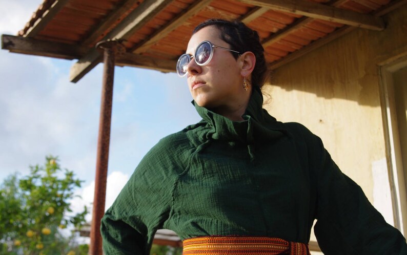 May include: A woman wearing a dark green, collared shirt with a ruffled neckline. She is wearing round, dark sunglasses and has a gold hoop earring in her right ear. She is standing under a wooden awning with a tiled roof.