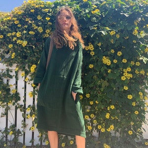 May include: A woman wearing a dark green, long-sleeved, loose-fitting dress with a pocket. She is standing in front of a white picket fence covered in green vines with yellow flowers.