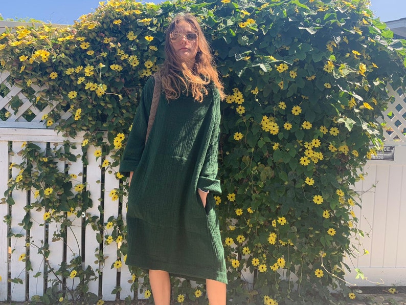May include: A woman wearing a dark green, long-sleeved, loose-fitting dress with a pocket. She is standing in front of a white picket fence covered in green vines with yellow flowers.