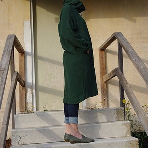May include: A woman wearing a dark green long-sleeved dress with a high collar, standing on a set of concrete steps. She is wearing blue jeans and dark green shoes.