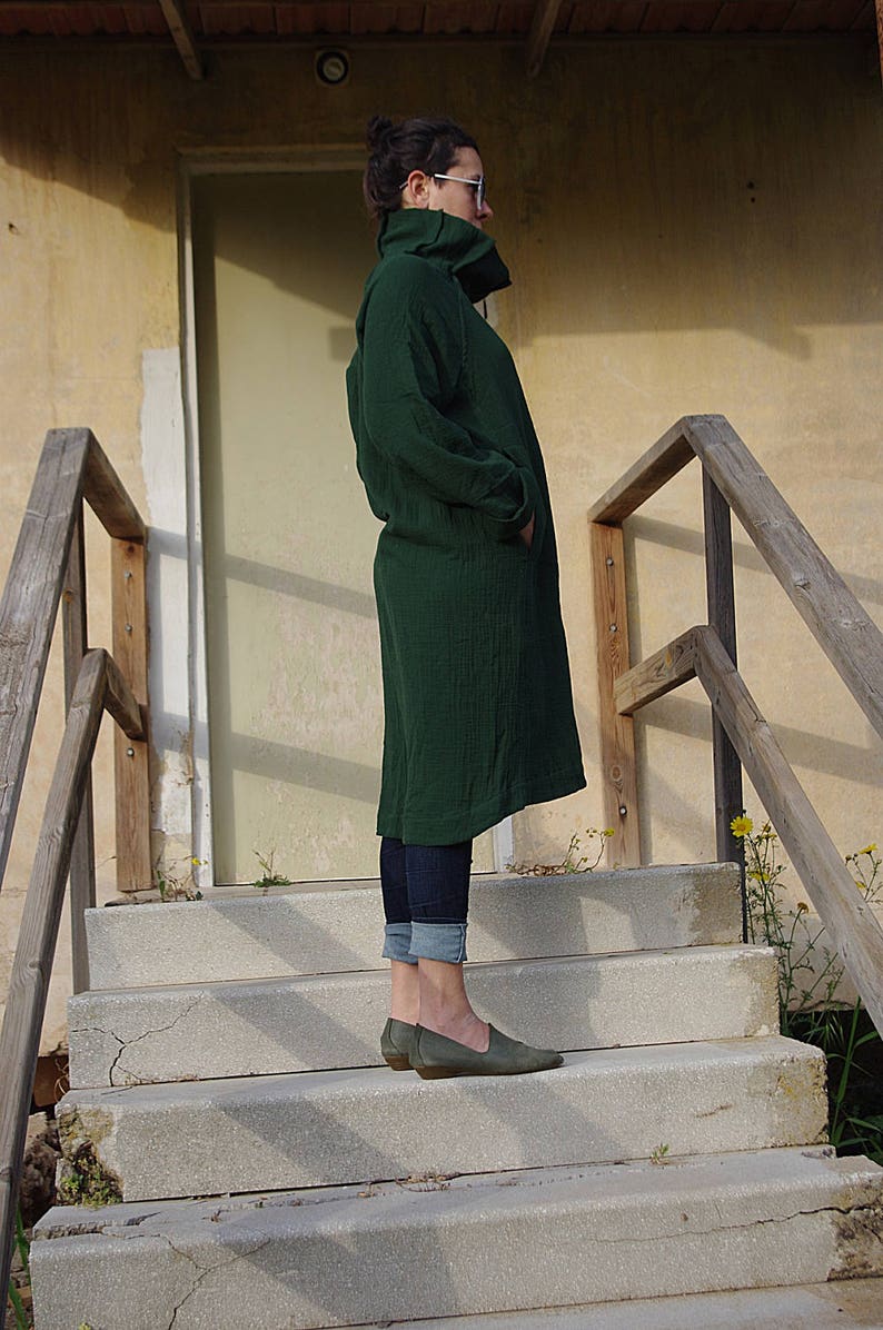 May include: A woman wearing a dark green long-sleeved dress with a high collar, standing on a set of concrete steps. She is wearing blue jeans and dark green shoes.