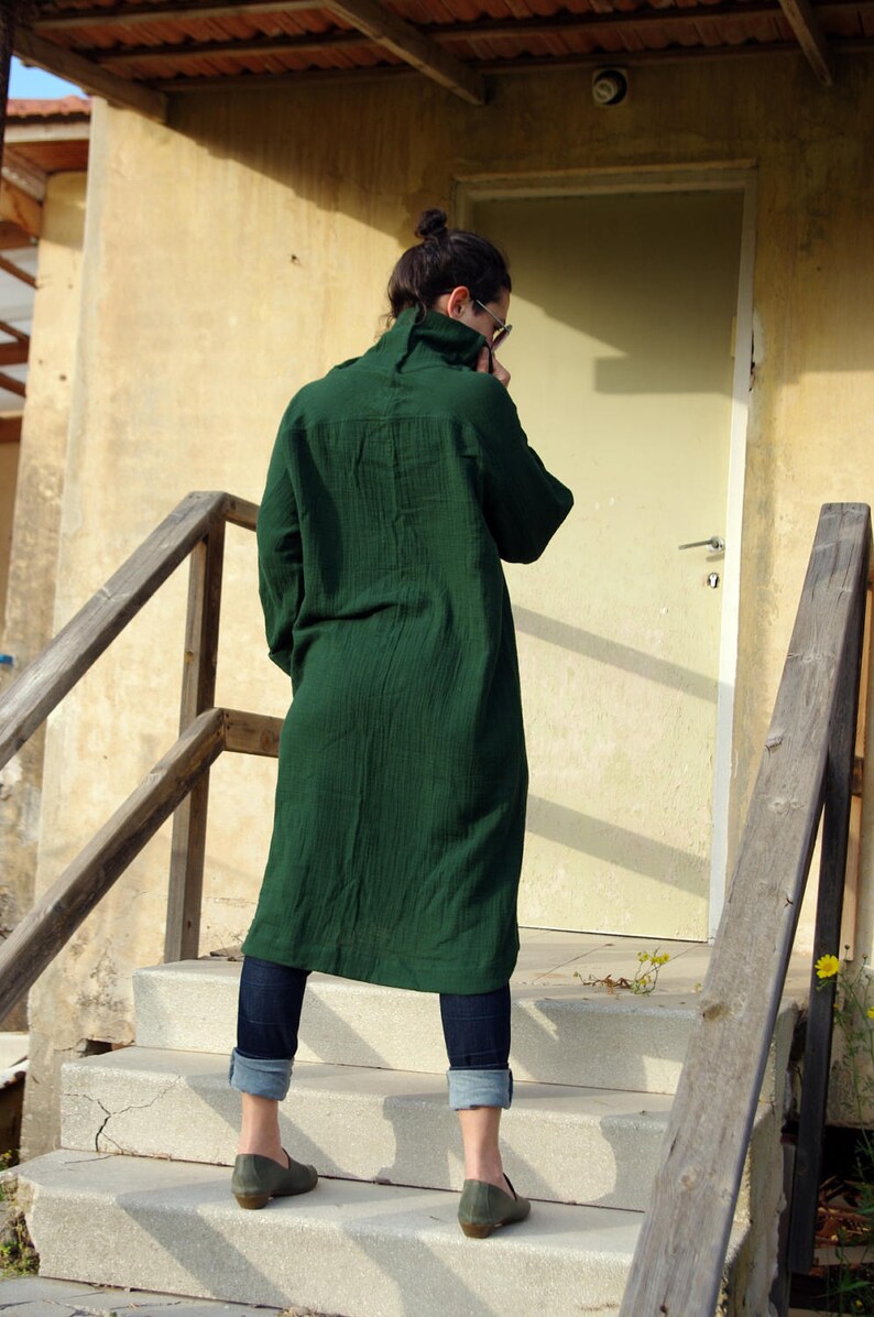 May include: A person wearing a long, dark green shirt and blue jeans stands on a set of concrete steps. The person is facing away from the camera and has their head down.
