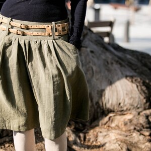 Könnte beinhalten: Ein grüner Leinenrock mit einem braunen Ledergürtel. Der Rock hat ein plissiertes Design und wird mit weißen Strumpfhosen getragen.