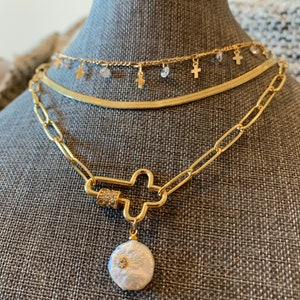 May include: A gold chain necklace with a large cross charm and a pearl pendant. The necklace is layered with two other gold chains, one with small cross charms and the other a simple chain.