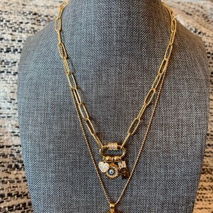 Pu&ograve; includere: Collana a strati in oro con un ciondolo che presenta un occhio malocchio, un cuore e la lettera "M". La collana ha anche un ciondolo con la parola "LOVE" circondata da strass.