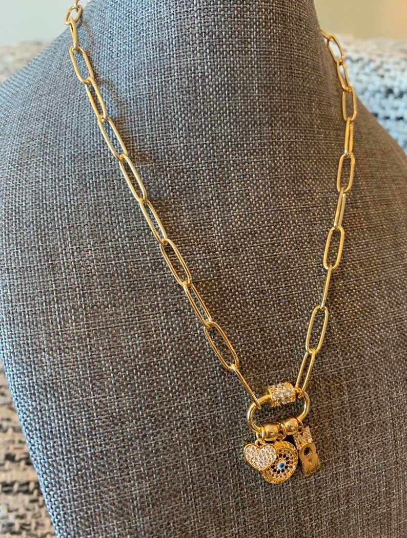 Pu&ograve; includere: Una collana a catena dorata con un ciondolo a forma di cuore, un occhio malvagio e un piccolo ciondolo con una lettera. La catena &egrave; in metallo dorato e ha una chiusura a moschettone.
