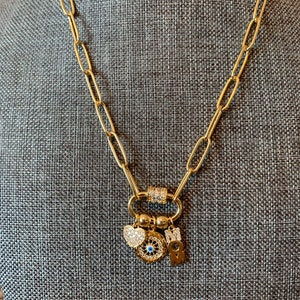Pu&ograve; includere: Collana a catena dorata con un ciondolo che presenta un occhio turco blu, un cuore e una lettera "M". I ciondoli sono attaccati a una chiusura dorata.