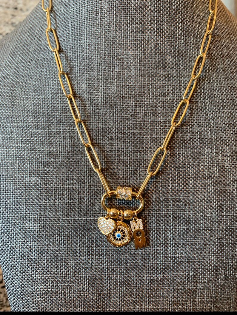 Pu&ograve; includere: Collana a catena dorata con un ciondolo che presenta un occhio turco blu, un cuore e una lettera "M". I ciondoli sono attaccati a una chiusura dorata.