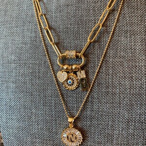 Pu&ograve; includere: Collana a strati in oro con un ciondolo a forma di cuore, occhio malocchio e ciondoli con lettere. Lo strato inferiore presenta un ciondolo rotondo con una lettera e accenti di cristallo.