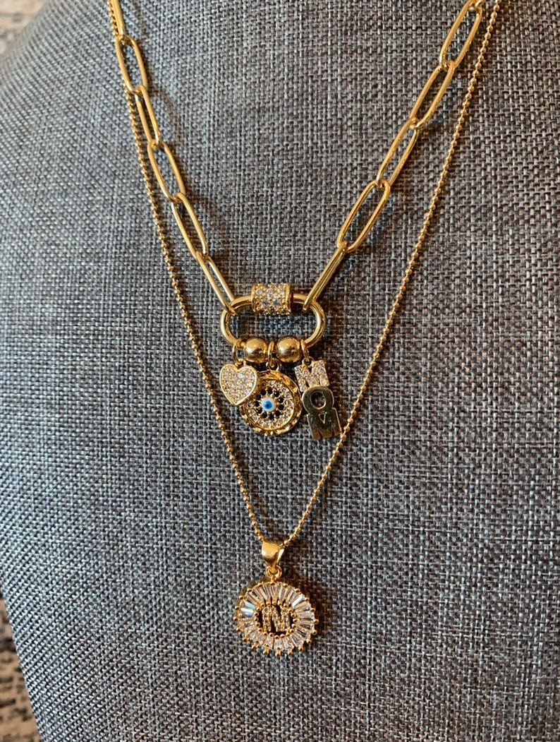 Pu&ograve; includere: Collana a strati in oro con un ciondolo a forma di cuore, occhio malocchio e ciondoli con lettere. Lo strato inferiore presenta un ciondolo rotondo con una lettera e accenti di cristallo.