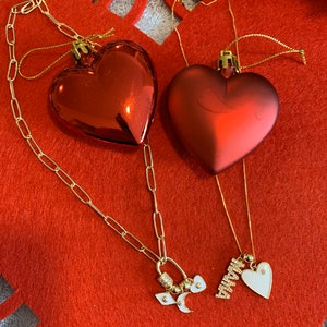 Pu&ograve; includere: Due collane d'oro con ciondoli a forma di cuore. Una collana ha un ciondolo con la parola "Mamma" e un piccolo cuore bianco. L'altra collana ha un ciondolo con una mezzaluna, una stella e un piccolo cuore bianco.