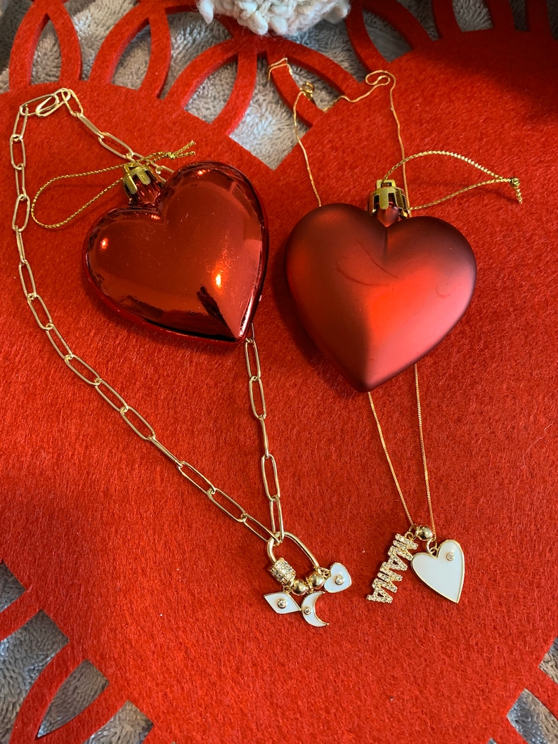 Pu&ograve; includere: Due collane d'oro con ciondoli a forma di cuore. Una collana ha un ciondolo con la parola "Mamma" e un piccolo cuore bianco. L'altra collana ha un ciondolo con una mezzaluna, una stella e un piccolo cuore bianco.