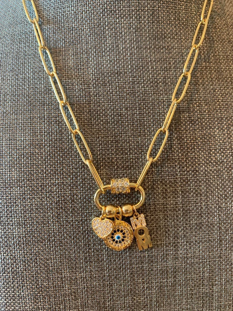 Pu&ograve; includere: Una collana a catena dorata con un ciondolo che presenta un occhio blu malocchio, un cuore e le lettere "MOM".