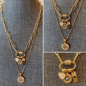 Pu&ograve; includere: Collana a strati in oro con un ciondolo a forma di occhio malocchio, un cuore e una lettera "M". La collana ha anche un ciondolo a forma di cerchio con le lettere "M" e "N" all'interno.
