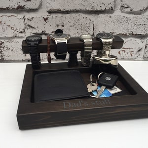 May include: A dark brown wooden tray with a compartment for a wallet, keys, and other small items. The tray has a raised section with four slots for watches and a small space for a bracelet. The tray is engraved with the words "Dad's stuff".