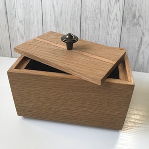 May include: A wooden box with a hinged lid, partially open. The box is made of light brown wood with a darker brown accent on the lid. A small, round, dark metal knob serves as a handle. The interior appears to be lined with black fabric.