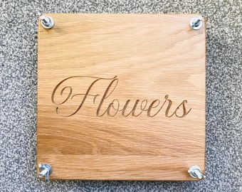 May include: A square wooden flower press with the word "Flowers" carved into the light brown wood. The press is held together with four metal bolts and wingnuts, one in each corner. The wood grain is visible.