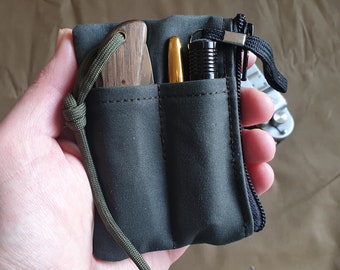 Può includere: Un organizer da tasca in tela verde oliva scuro con due scomparti verticali e una cerniera laterale. Gli scomparti contengono un coltello pieghevole con manico in legno e una penna dorata, mentre una torcia nera è fissata sul lato con un cordino.