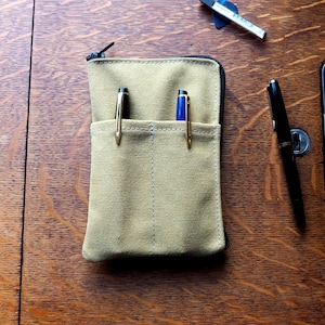 Può includere: Un astuccio portapenne in tela color sabbia con chiusura a zip nera e due tasche frontali, ciascuna contenente una penna. L'astuccio è rettangolare con bordi cuciti. A destra si trova una penna stilografica nera e in alto un piccolo righello in metallo.