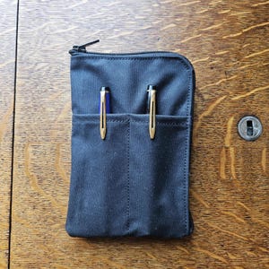 May include: A black fabric pen case with a top zipper closure and two exterior front pockets. Each pocket contains a pen with a gold-colored clip. The case has a rectangular shape with rounded top corners and visible stitching along the edges.