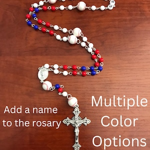 May include: Baseball themed rosary with white, red, and blue beads. The rosary has a silver crucifix and a baseball charm. The text "JOSEPH" is spelled out with letter beads. The text "Add a name to the rosary" and "Multiple Color Options" is also visible in the image.