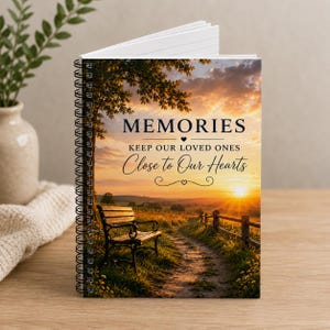 May include: A spiral-bound notebook featuring a sunset landscape on the cover with a wooden bench along a path. The text reads: MEMORIES KEEP OUR LOVED ONES Close to Our Hearts. The cover displays a golden sky, green fields, and a wooden fence.