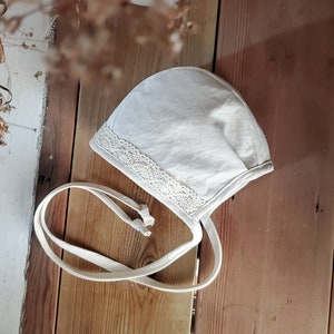 Könnte beinhalten: Ein weißer Baby-Mützchen aus Leinen mit einer Spitzenborte und einer weißen Schleife. Die Mütze liegt auf einer Holzoberfläche.