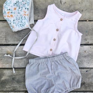 Peut inclure: Un ensemble de vêtements pour bébé comprenant un haut sans manches rose clair avec des boutons en bois, un bloomer à carreaux gris et un bonnet à imprimé floral. Le bonnet a un motif floral bleu clair, orange et blanc.