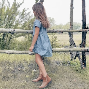 May include: A young person wearing a light blue denim dress and brown ankle boots stands in front of a wooden fence. The person is looking to the left of the frame.