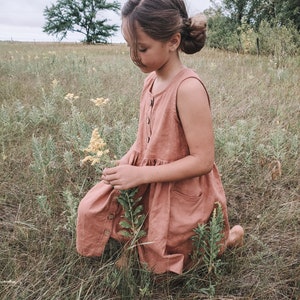 May include: A young girl in a dusty pink sleeveless dress with buttons down the front is kneeling in a field of tall grass. She is holding a sprig of yellow wildflowers.