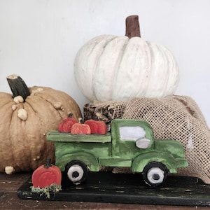 May include: A green wooden toy truck carrying small red pumpkins, with a large white pumpkin and a tan pumpkin in the background. The truck is on a black surface, with a burlap sack and wood in the background. Autumn decor.