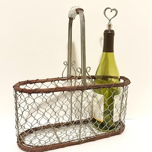 May include: An oval wire wine basket with a brown rim and handle, holding a green wine bottle. A heart-shaped bottle stopper is in the bottle. The handle has a white ceramic accent.