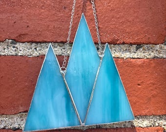 May include: A stained glass mountain range in shades of blue and turquoise, with silver-colored metal edging and chain for hanging. The glass has a textured appearance, and the piece is mounted on a red brick wall.