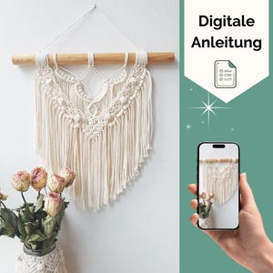 Könnte beinhalten: Cremefarbener Makramee-Wandbehang mit einem Holzdübel. Das Design weist komplizierte Knoten und Fransen auf. Das Bild zeigt auch ein Smartphone, das das gleiche Makramee-Stück anzeigt, und den Text "Digitale Anleitung".