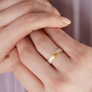 May include: A silver band ring featuring a gold-toned sunburst or fan-shaped engraving on the outer surface. The ring has a smooth, flat profile and a polished finish, with the metallic gold detail contrasting against the silver-colored metal band.