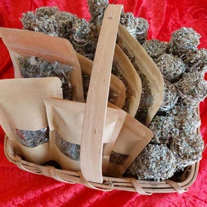 Può includere: Un cesto di legno pieno di mazzi di erbe secche avvolti in sacchetti di carta marrone. Le erbe sono di colore verde chiaro e grigio.