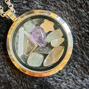 Puede incluir: Collar con colgante de medallón plateado lleno de piedras preciosas de colores. El colgante redondo tiene una parte delantera de cristal transparente, que muestra piedras moradas, verdes y beige. La cadena del collar también es plateada.