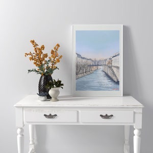 Peut inclure: Une console blanche avec deux tiroirs et des poign&eacute;es de tiroir argent&eacute;es. La table a une finition blanchie &agrave; la chaux et est d&eacute;cor&eacute;e d'un tableau encadr&eacute;e d'un paysage urbain parisien avec une rivi&egrave;re et un pont.