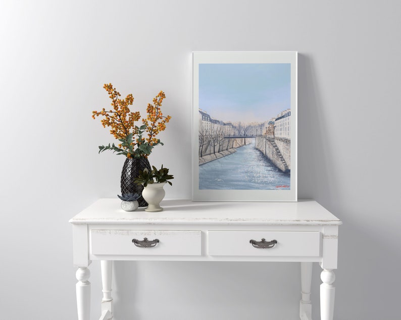 Peut inclure: Une console blanche avec deux tiroirs et des poign&eacute;es de tiroir argent&eacute;es. La table a une finition blanchie &agrave; la chaux et est d&eacute;cor&eacute;e d'un tableau encadr&eacute;e d'un paysage urbain parisien avec une rivi&egrave;re et un pont.