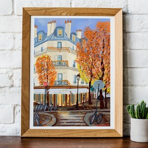 May include: A framed painting of a Parisian street scene with a cafe, bicycles, and trees in autumn colors. The building is white with blue accents and has a sign that reads "Le Cafe de la Paix".