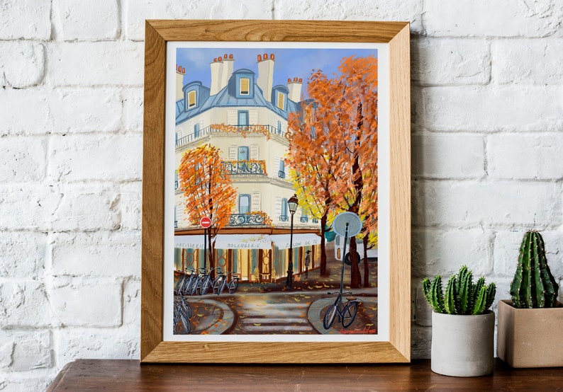 May include: A framed painting of a Parisian street scene with a cafe, bicycles, and trees in autumn colors. The building is white with blue accents and has a sign that reads "Le Cafe de la Paix".