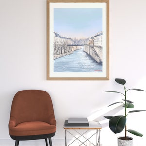 Peut inclure: Une peinture &agrave; l'aquarelle encadr&eacute;e d'un paysage urbain parisien avec une rivi&egrave;re qui coule au centre. Les b&acirc;timents sont blancs avec des toits bruns et des arbres bordent les rives de la rivi&egrave;re.