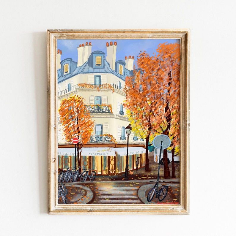 May include: A painting of a Parisian street scene with a building, trees, and bicycles. The building is white with blue trim and has a sign that says "Boulangerie". The trees are orange and yellow and the bicycles are parked on the sidewalk. The painting is framed in a light wood frame.