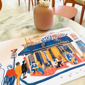 Puede incluir: Una ilustraci&oacute;n colorida de un caf&eacute; parisino con gente sentada afuera. La ilustraci&oacute;n est&aacute; impresa en una tarjeta blanca y est&aacute; sobre una mesa de m&aacute;rmol con un jarr&oacute;n rosa con flores blancas.