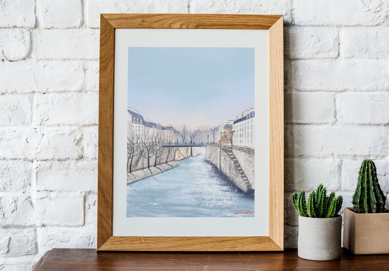 Peut inclure: Une peinture &agrave; l'aquarelle encadr&eacute;e d'une sc&egrave;ne de rue parisienne avec un pont au-dessus d'un canal. Les b&acirc;timents sont blancs et l'eau est bleue. Le ciel est bleu clair.