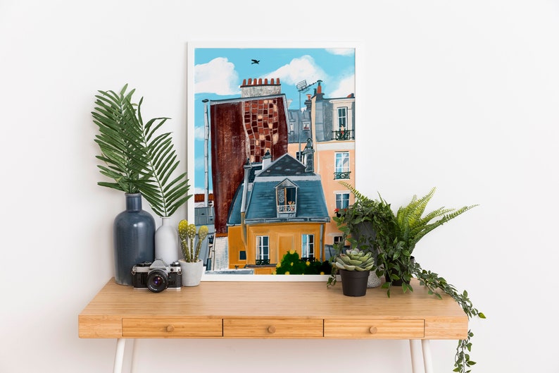 May include: A framed illustration of a colourful cityscape with buildings in shades of orange, blue, and brown. The buildings have windows and balconies, and there is a small aeroplane flying in the sky above.