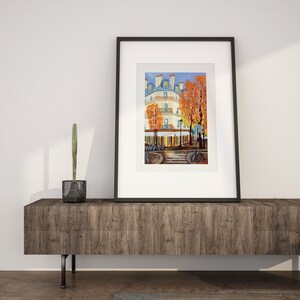 May include: A framed painting of a Parisian street scene with a white building, trees with orange leaves, and people walking by. The painting is in a black frame and is sitting on a wooden shelf.