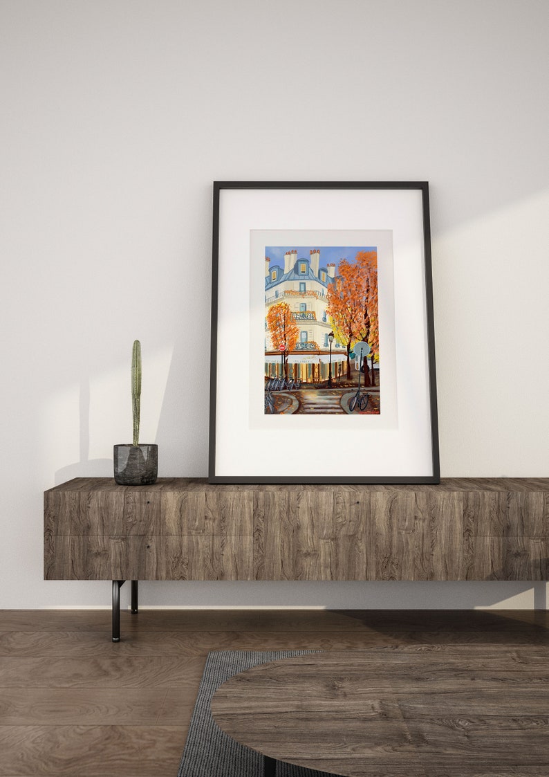 May include: A framed painting of a Parisian street scene with a white building, trees with orange leaves, and people walking by. The painting is in a black frame and is sitting on a wooden shelf.